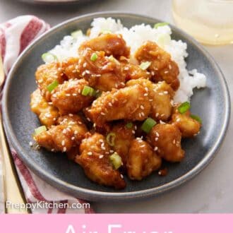 Pinterest graphic of a plate of rice and air fryer orange chicken garnished with sesame seeds and green onions. Another plate in the back along with a drink.