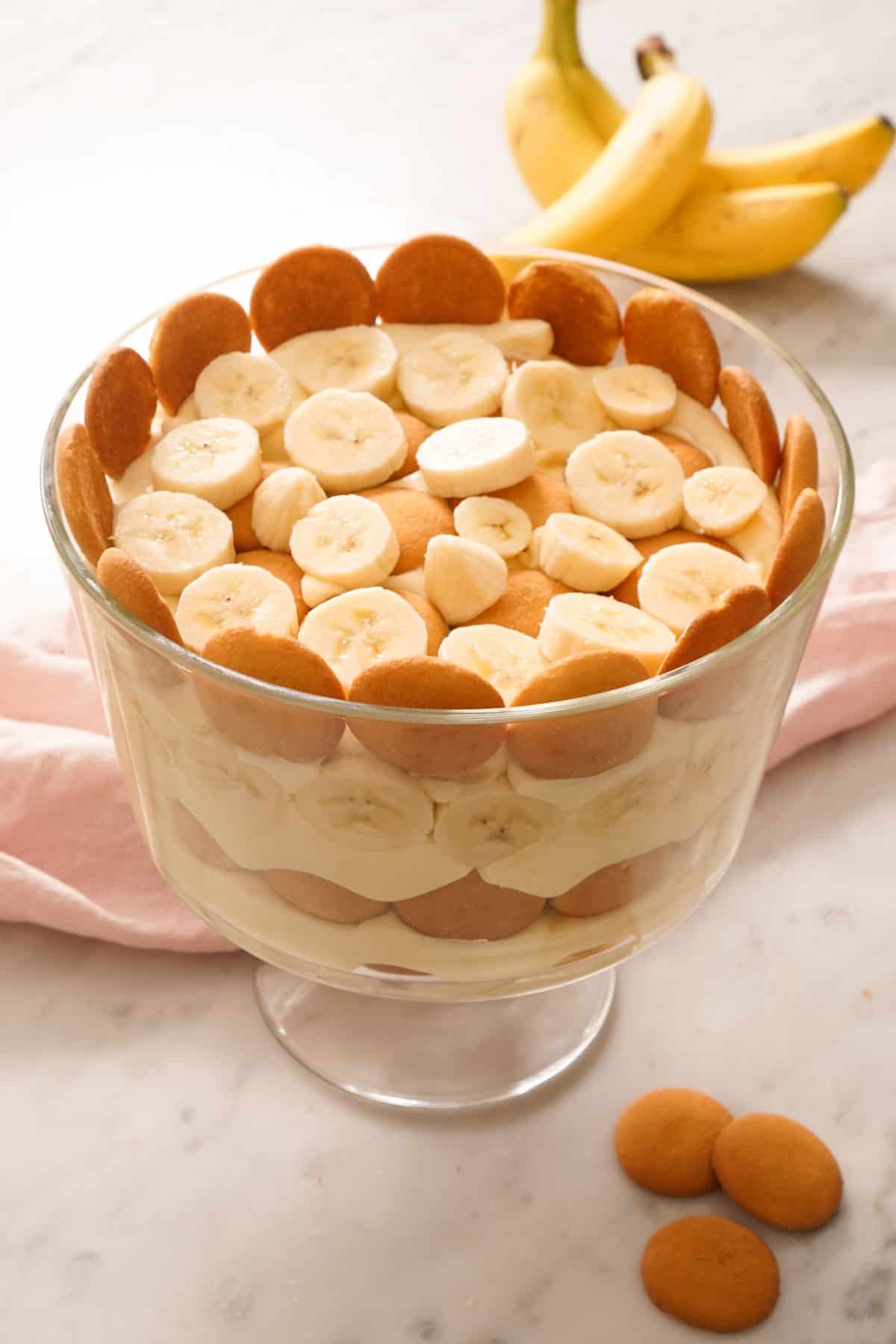Banana pudding in a trifle dish. Three bananas in the background with some crackers in the front.