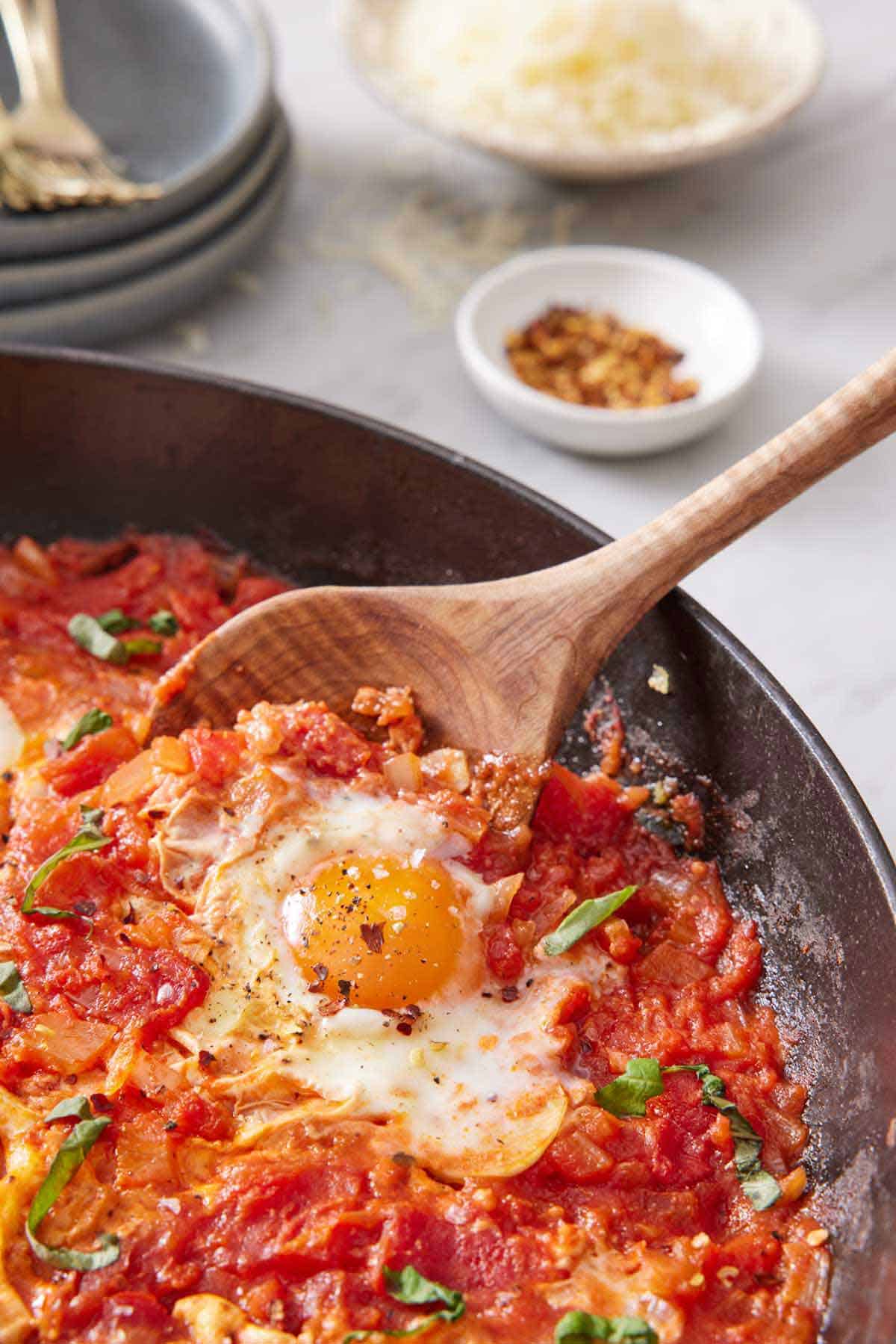 A spoon scooping up eggs in purgatory in a skillet. Bowl of red pepper flakes and shredded cheese in the background.