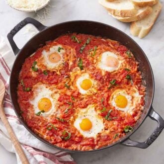 An overhead view of a skillet of eggs in purgatory. A wooden spoon and some sliced bread beside it.