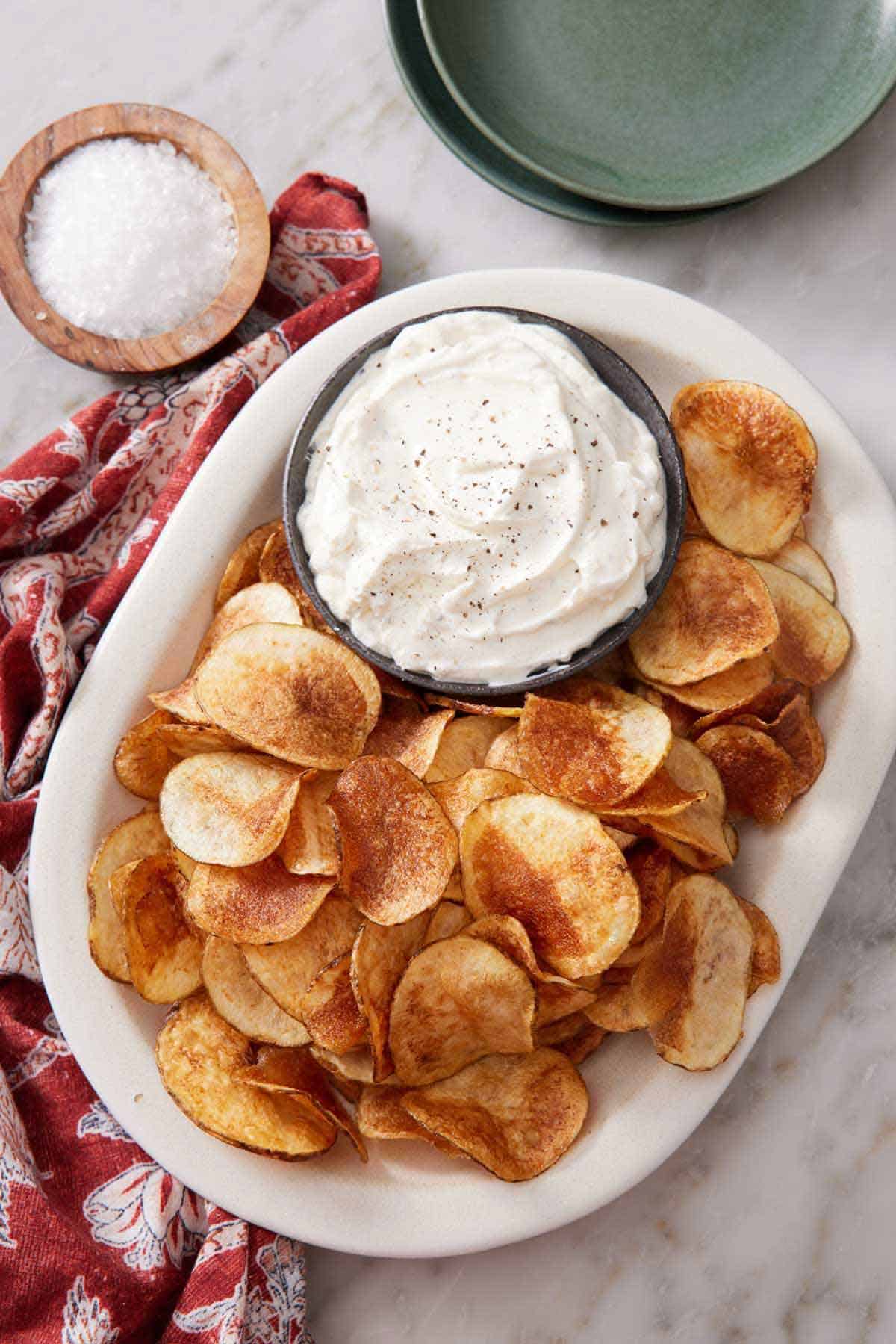 Overhead view of a platter of homemade potato chips with a bowl of dip. A small bowl of salt and some plates on the side.