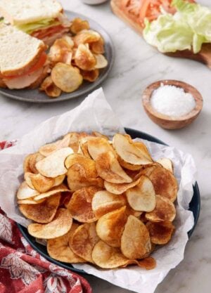 A plate lined with parchment paper topped with homemade potato chips. A plate with a cut sandwich with chips in the back along with a bowl of salt and sandwich ingredients.