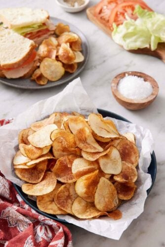 A plate lined with parchment paper topped with homemade potato chips. A plate with a cut sandwich with chips in the back along with a bowl of salt and sandwich ingredients.