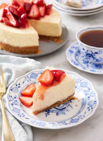 A plate with a slice of Instant Pot cheesecake topped with chopped strawberries. A mug of tea and the rest of the cheesecake in the background.