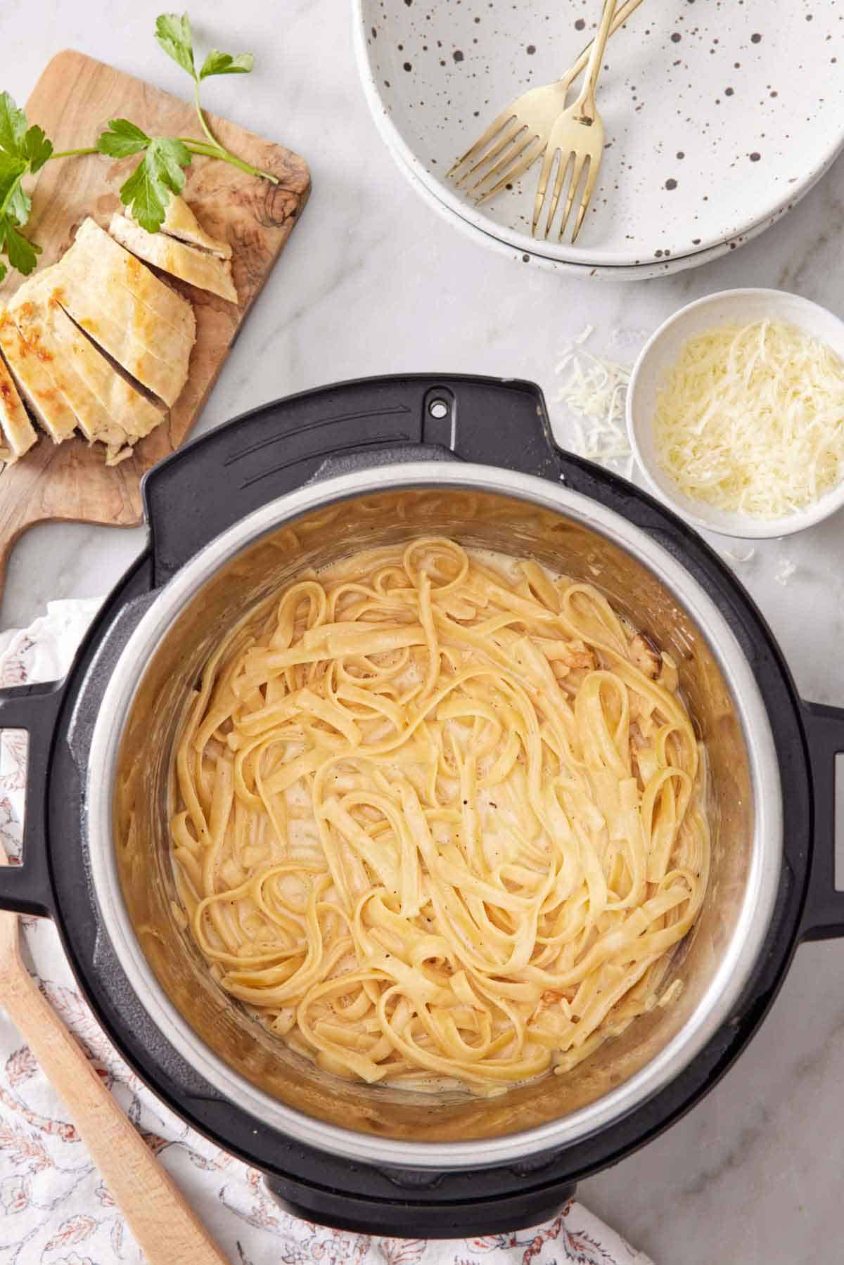 Overhead view of chicken alfredo in the Instant Pot. A stack of plates, forks, a bowl of cheese, and sliced bread with parsley on the side.