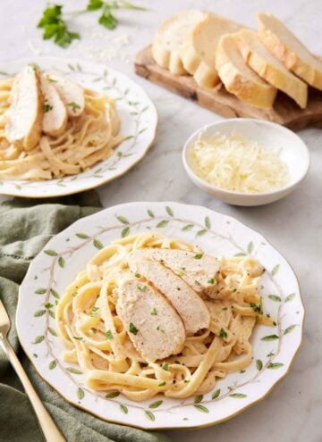 A plate with a serving of Instant Pot chicken alfredo with a second plate in the back along with a bowl of cheese and sliced bread.