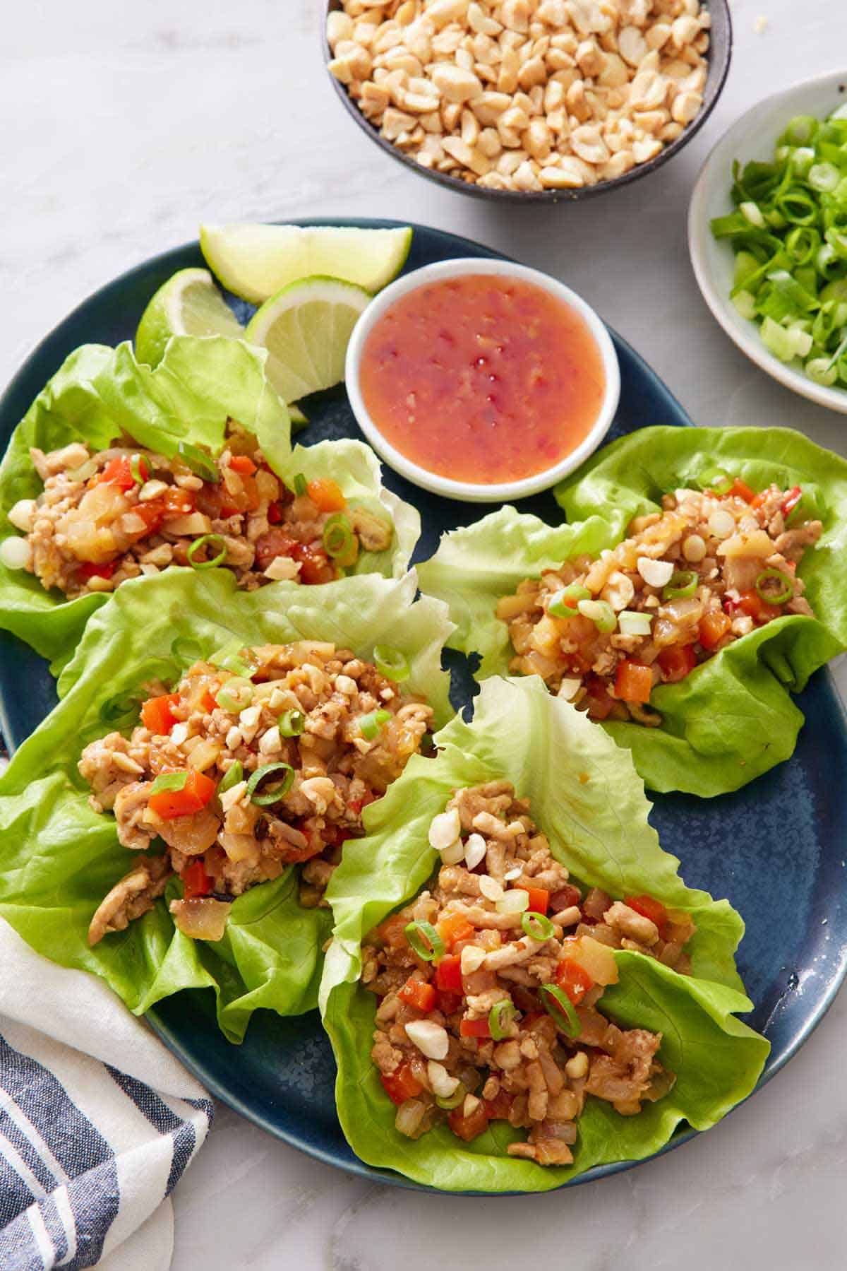 A platter of lettuce wraps with a sweet chili sauce in a bowl along with lime wedges. A bowl of peanuts and green onions off on the side.