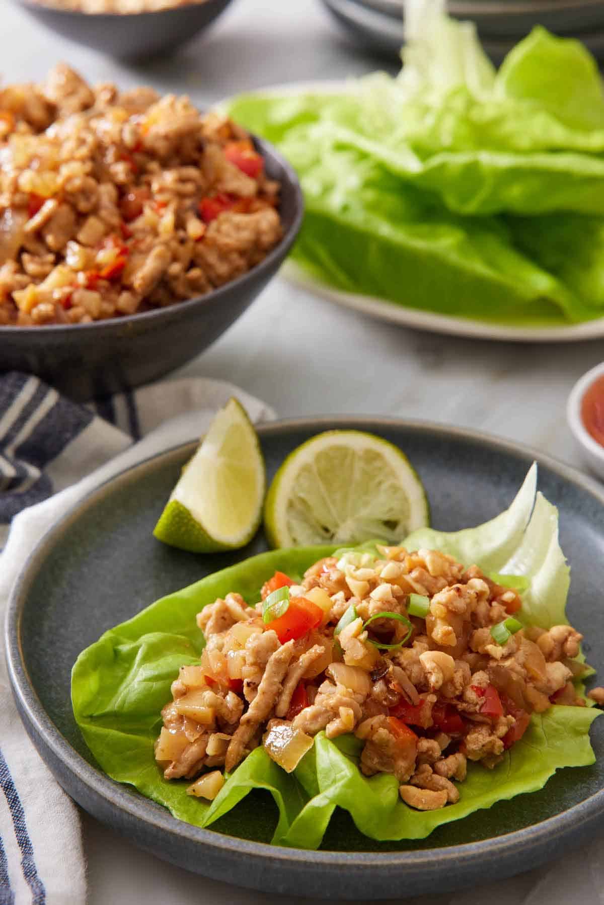 A plate with a serving of lettuce wrap with lime wedges. A bowl of filling and plate of lettuce in the back.