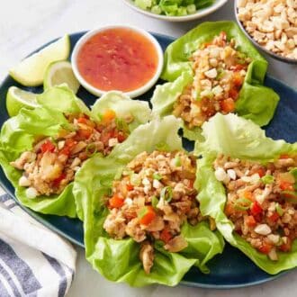 A platter of four lettuce wraps with a bowl sweet chili sauce and lime wedges. Bowl of peanuts in the back.