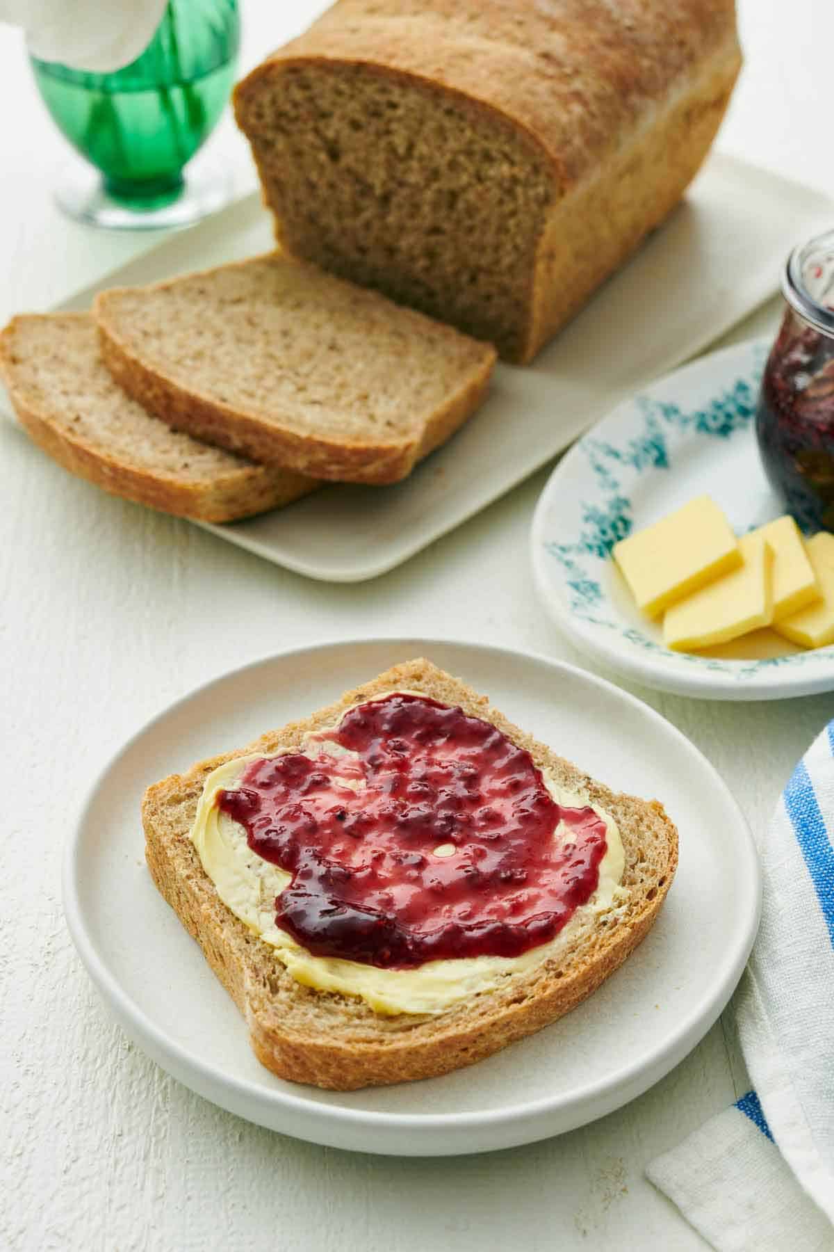 A plate with a slice of rye bread with peanut butter and jam spread on top. Rest of the loaf in the background with two more slices cut.