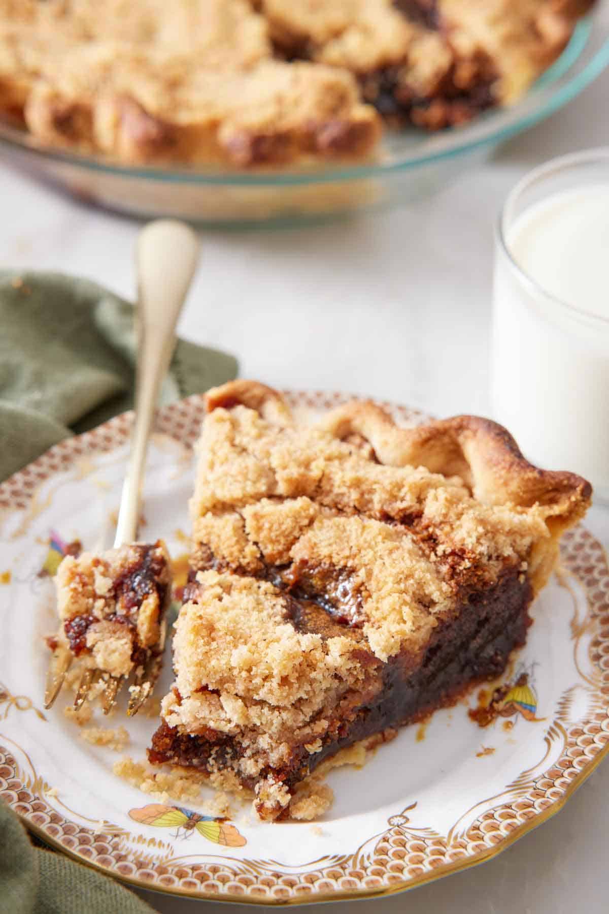 A plate with a slice of shoofly pie with the tip of the slice on the fork beside it. A glass of milk and the rest of the pie behind it.