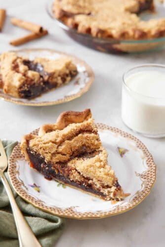 A plate with a slice of shoofly pie with a glass of milk, another plated serving, and the rest of the pie in the background.