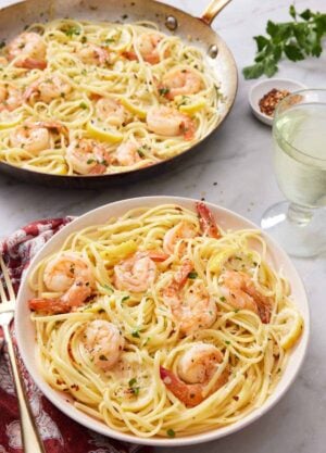 A plate with shrimp linguine with a glass of wine and skillet with more shrimp linguine in the back.