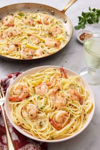 A plate with shrimp linguine with a glass of wine and skillet with more shrimp linguine in the back.