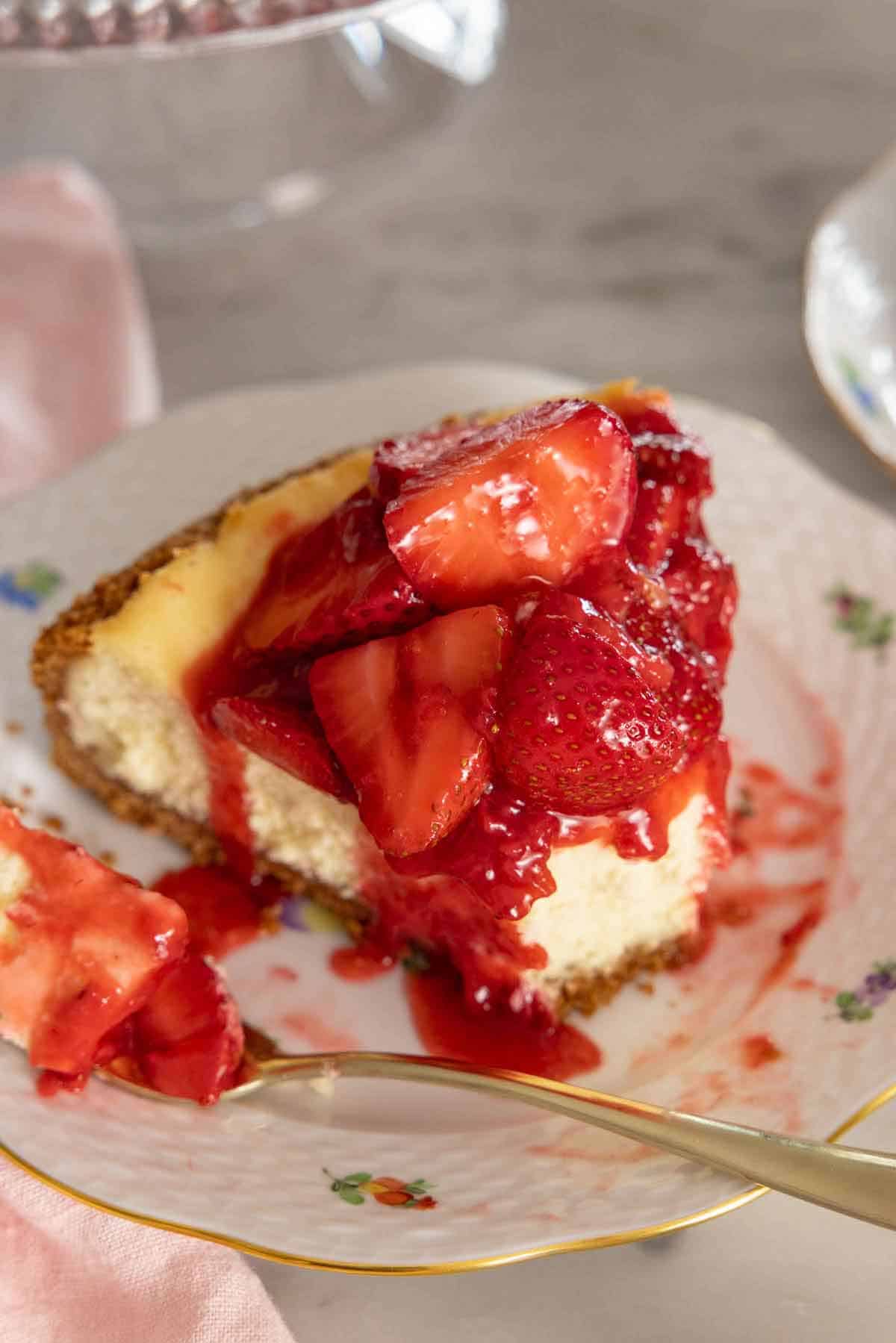 A slice of strawberry cheesecake on a plate with the tip of the slice on a fork beside it.