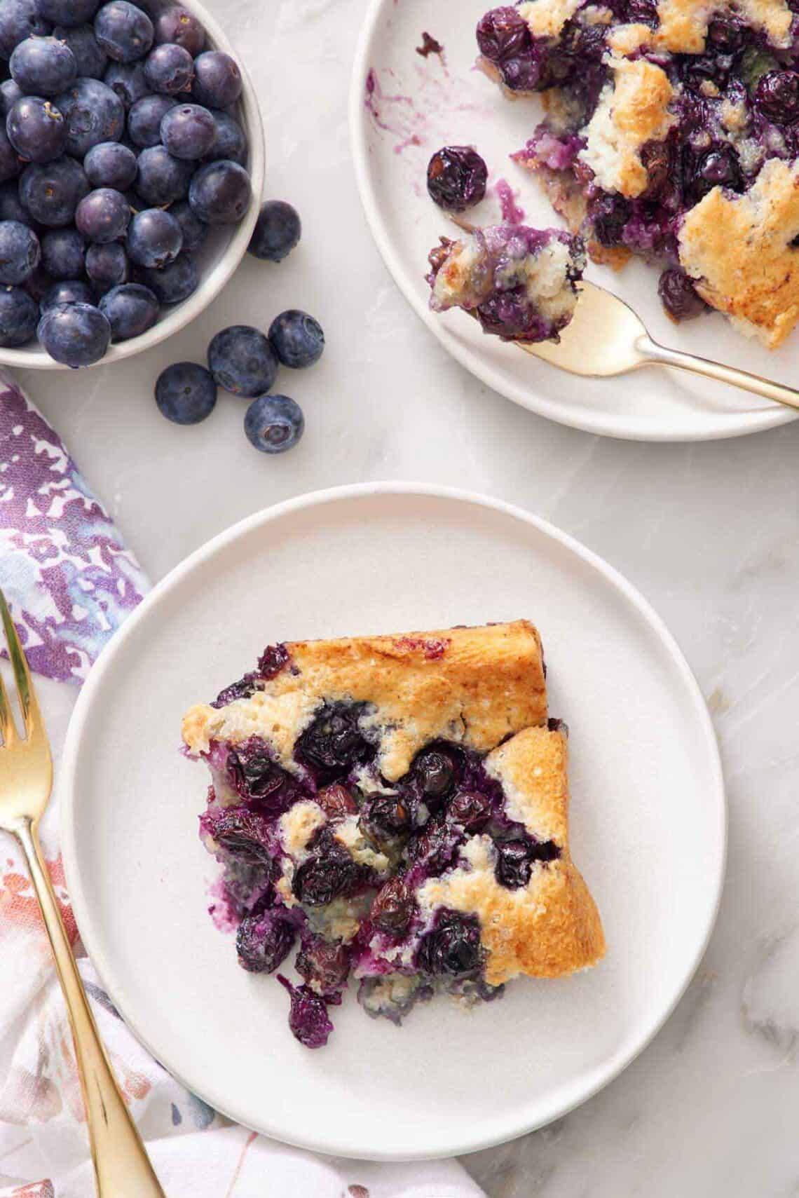 Overhead view of a slice of blueberry cobbler on a plate with another plate off to the side with a fork. A bowl of blueberries on the side.