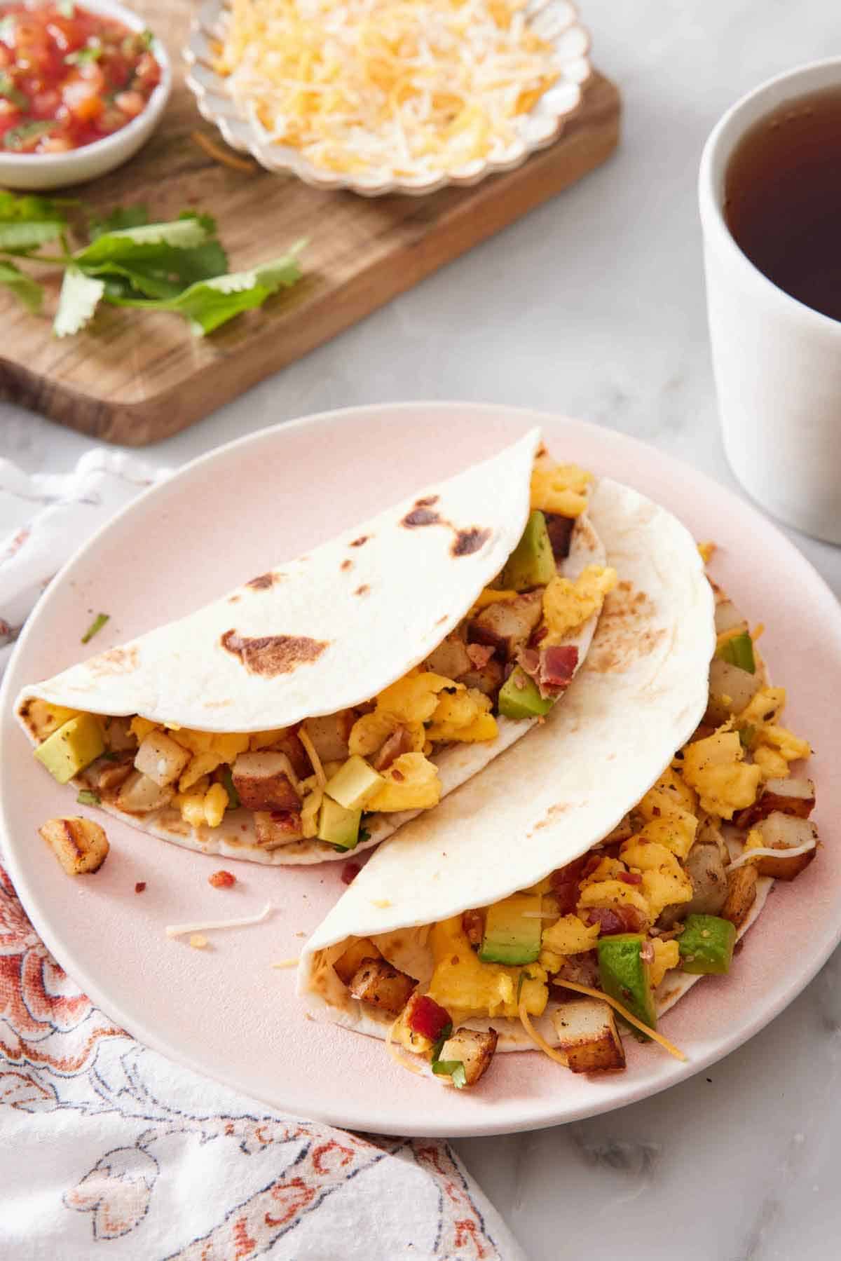 A plate with two breakfast tacos. Toppings and a mug of coffee in the background.