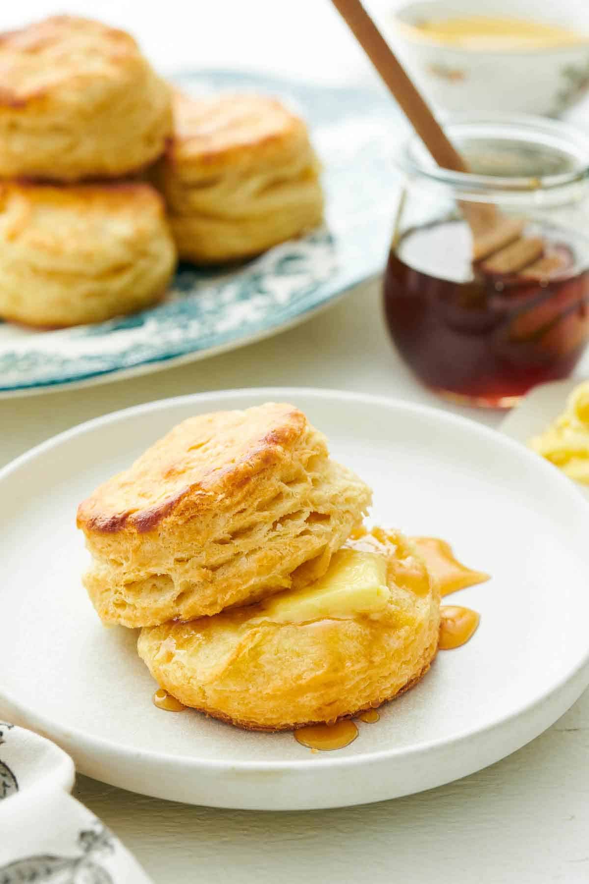 A plate with buttermilk biscuits with some honey and butter. A jar of honey in the background and more biscuits.