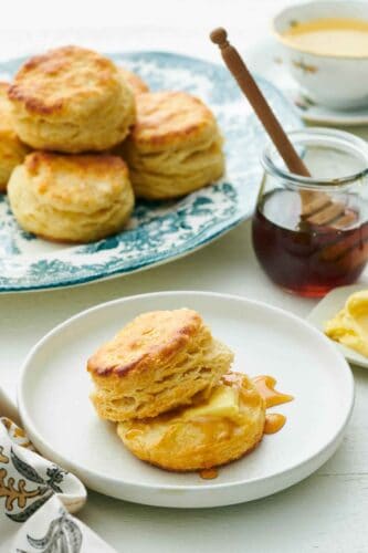 A plate with buttermilk biscuits with some honey and butter. A jar of honey and platter of biscuits in the background.