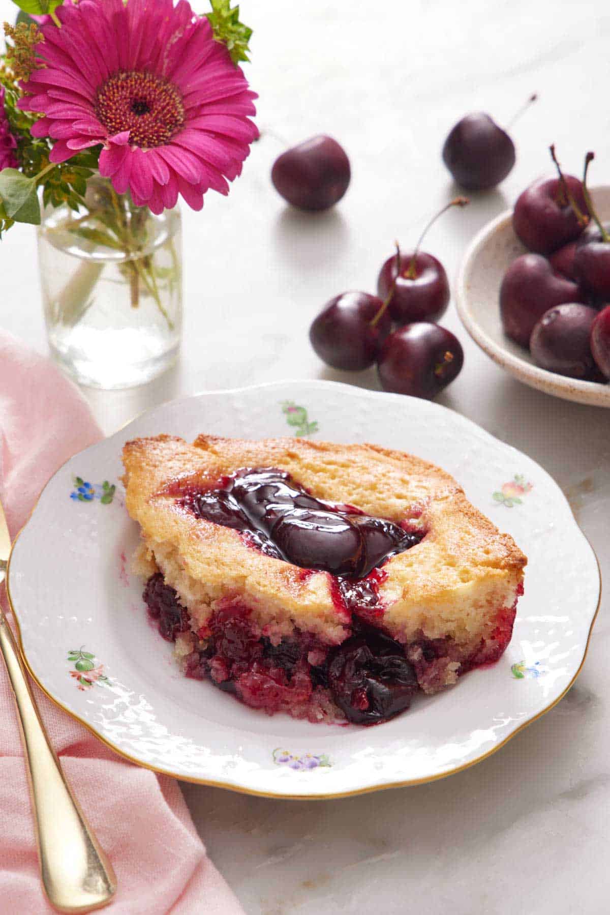 A plate with a serving of cherry cobbler with a small vase of flowers and cherries in the background.