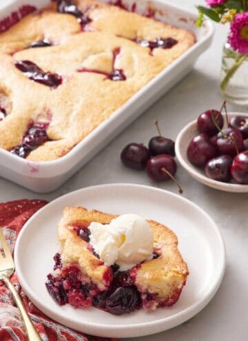 A plate with a serving of cherry cobbler with the rest of the cobbler in the background with a bowl of cherries.