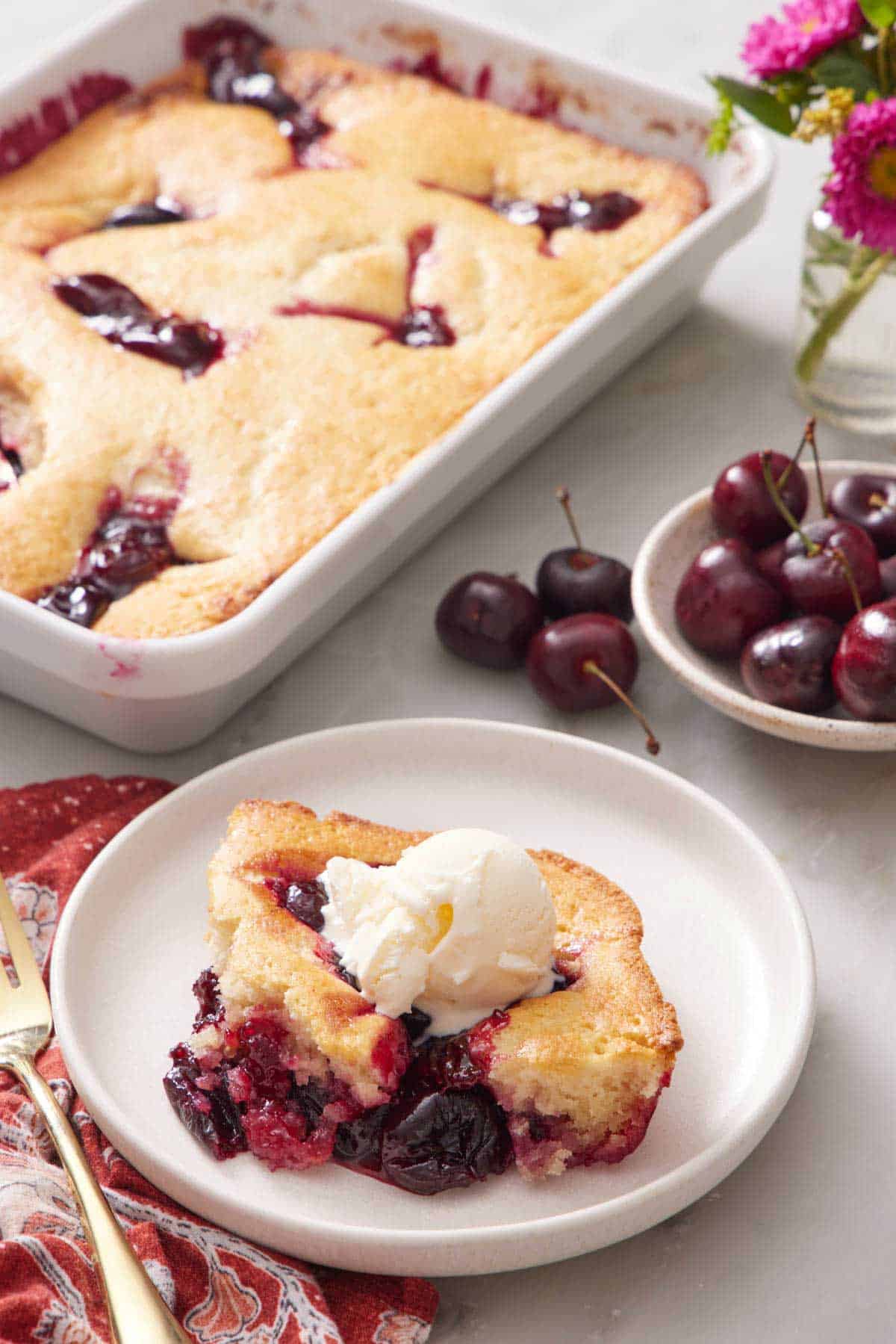 A plate with a serving of cherry cobbler with the rest of the cobbler in the background with a bowl of cherries.