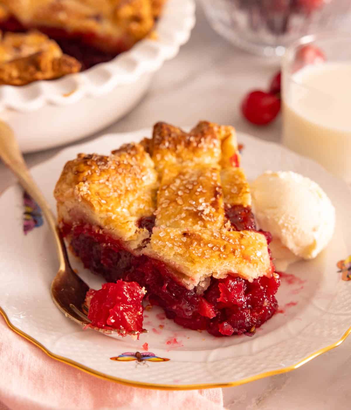A slice of cherry pie with a fork and scoop of ice cream beside it.