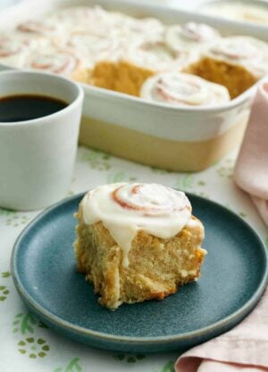 A plate with a cinnamon roll. A baking dish with more rolls and a mug of coffee in the back.