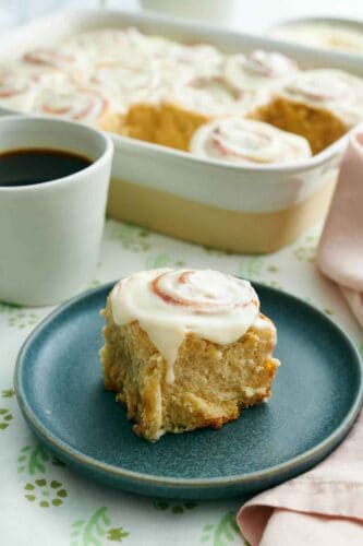 A plate with a cinnamon roll. A baking dish with more rolls and a mug of coffee in the back.
