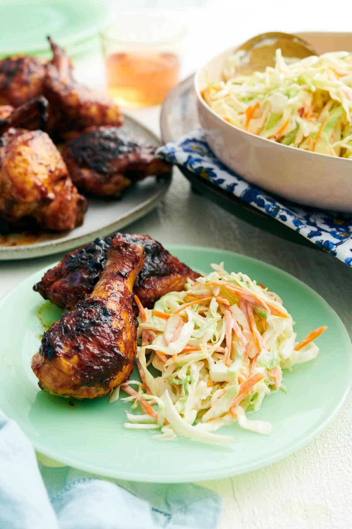 A plate with coleslaw and grilled chicken drumsticks. More chicken and coleslaw in the background.
