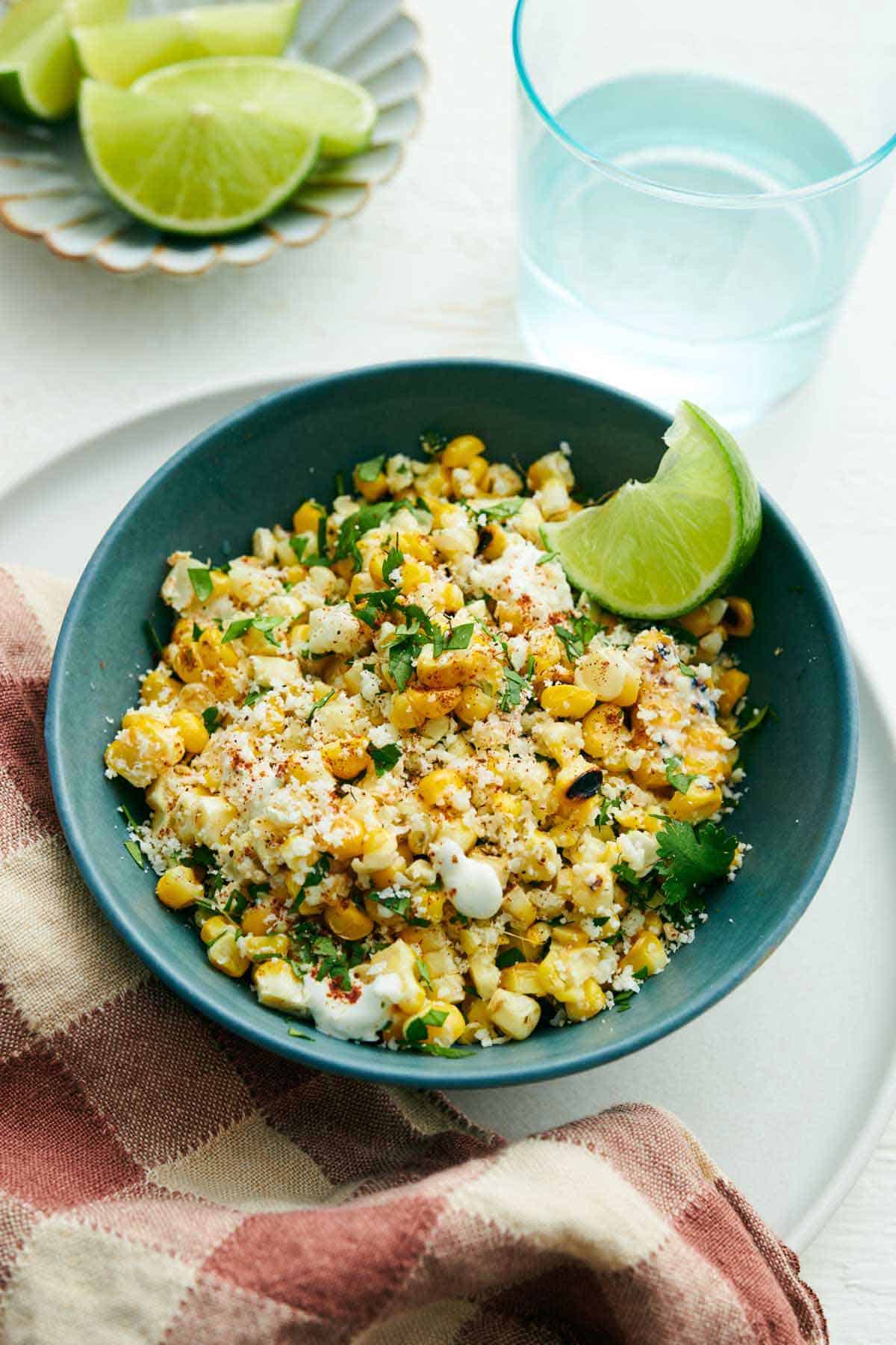 A bowl of elote corn topped with cilantro with a lime wedge. A glass of water and plate of more limes in the background.