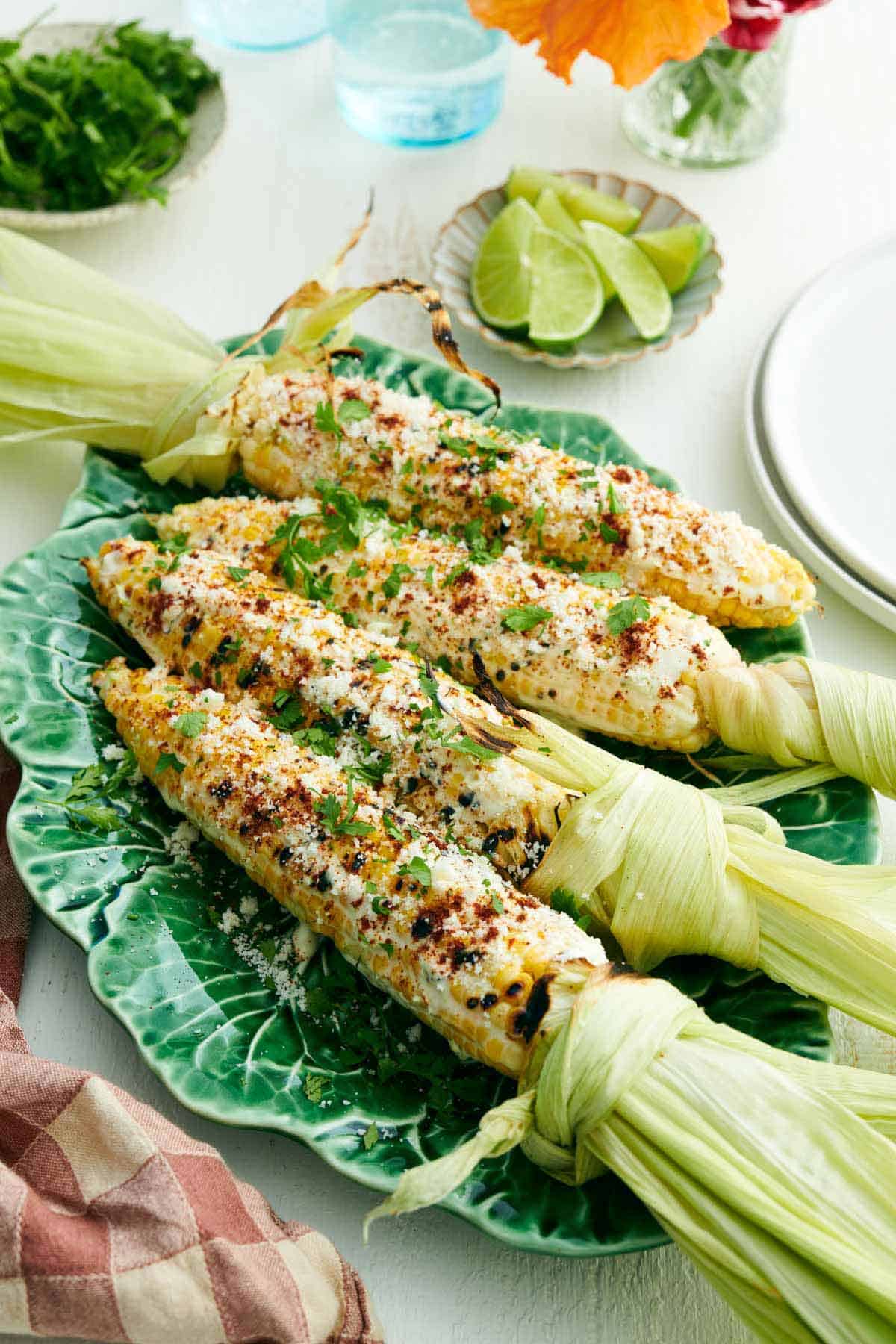 A platter with four elotes with cilantro sprinkled on top. Plate of limes and cilantro in the back.