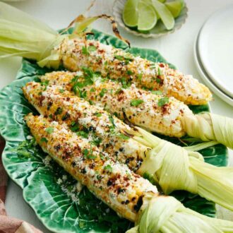 Pinterest graphic of a platter with four elotes with cilantro sprinkled on top. Plate of limes and cilantro in the back.