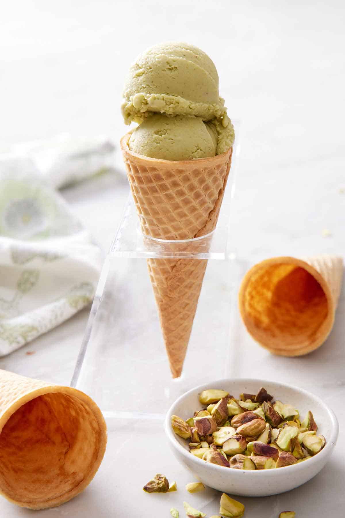 An ice cream cone with two scoops of pistachio ice cream. A bowl of pistachios in front with two cones scattered.