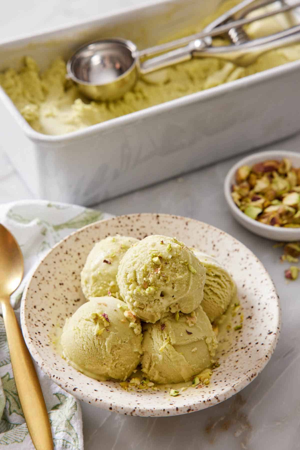 A plate with multiple scoops of pistachio ice cream with more ice cream in the background in a container with an ice cream scoop.