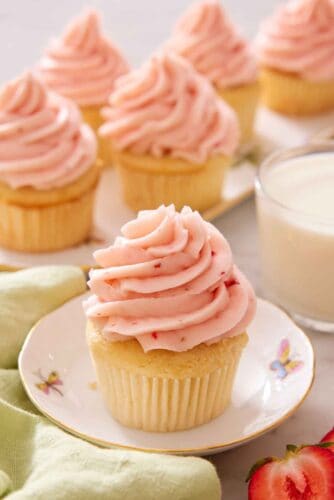 A cupcake topped with strawberry frosting with more topped cupcakes in the background along with a glass of milk.