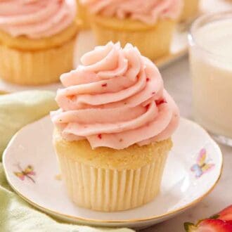 A plate with a cupcake topped with strawberry frosting with a glass of milk and more cupcakes in the background.