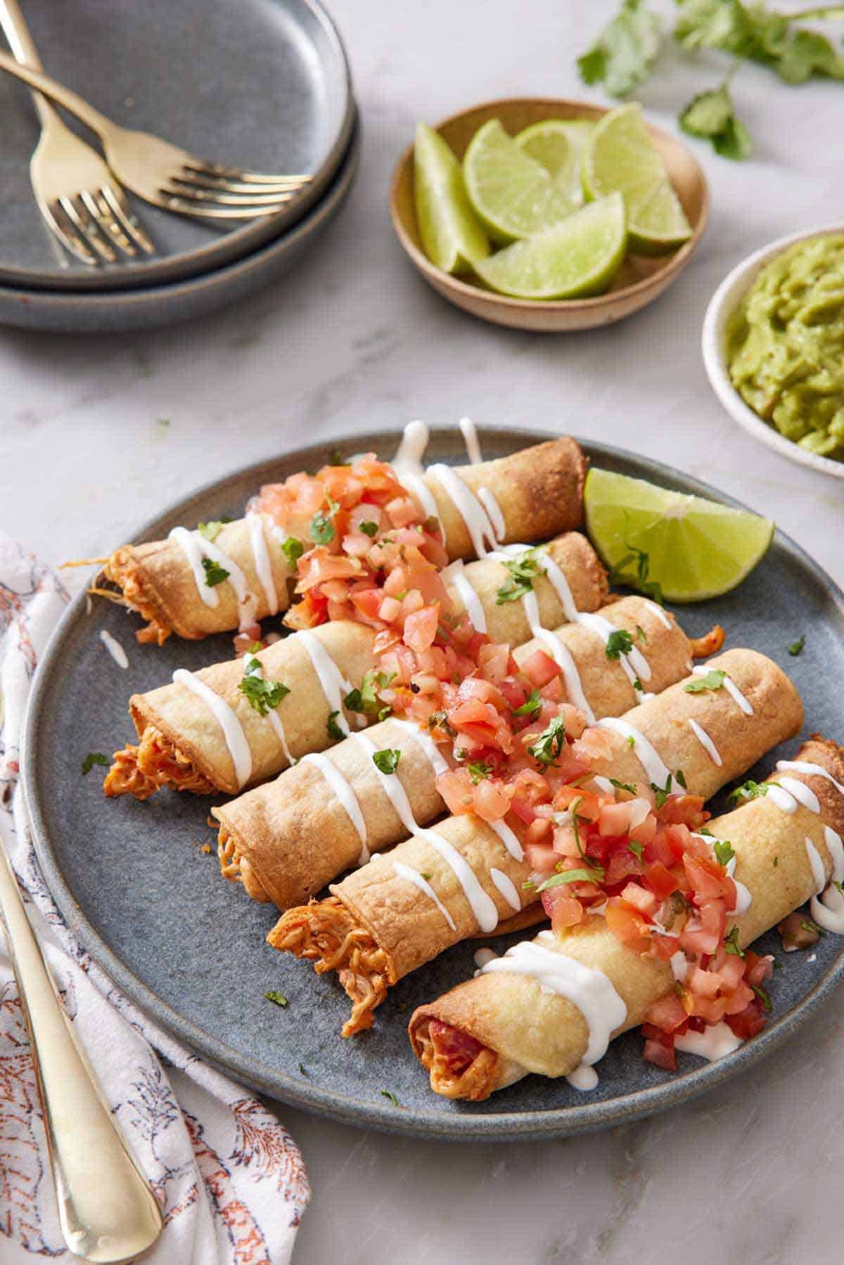 A plate of a lime wedge and air fryer taquitos topped with sour cream and salsa. More lime wedges in the background along with plates, forks, and guacamole.