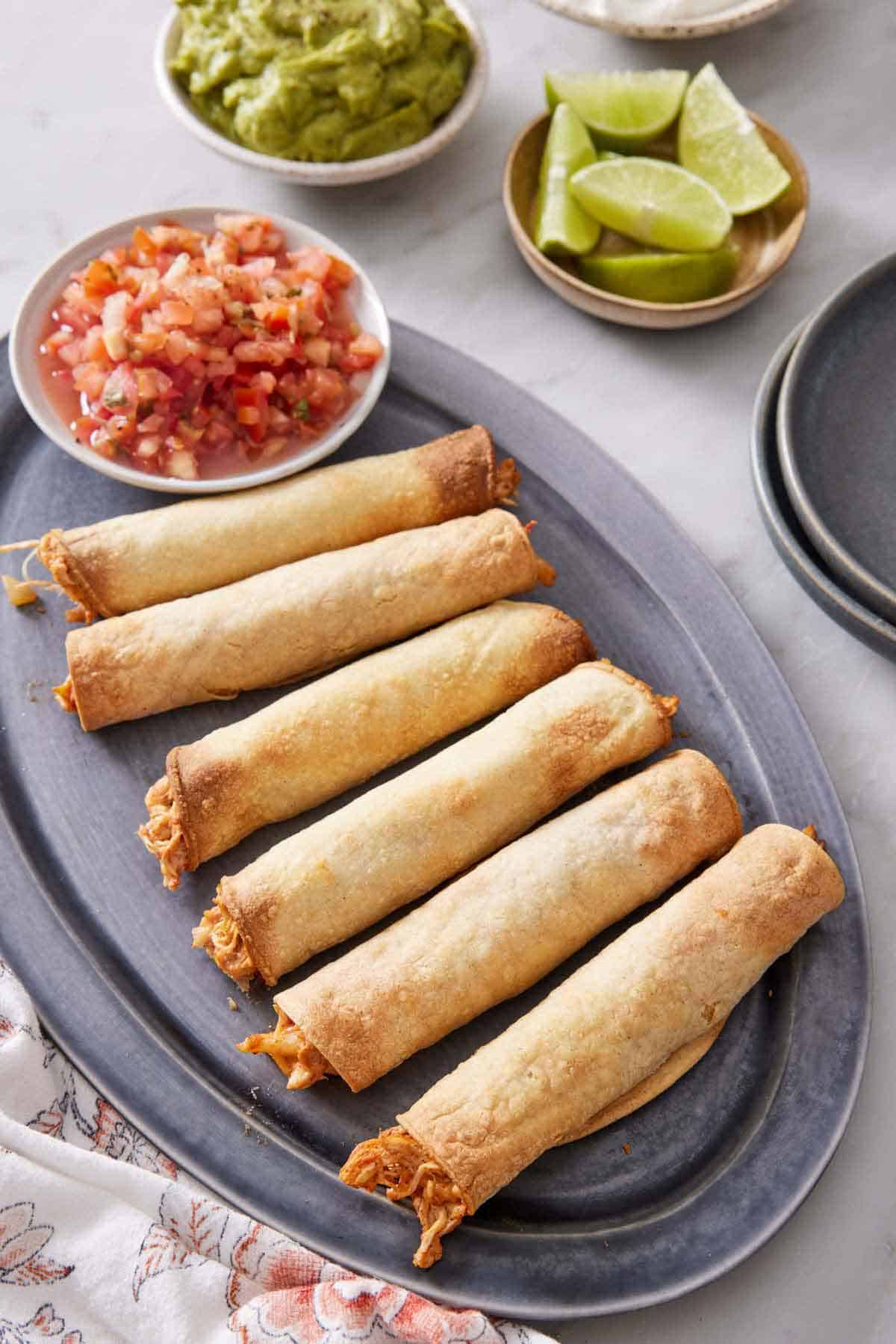 A platter of six air fryer taquitos with a bowl of salsa. Guacamole and lime wedges in the back.