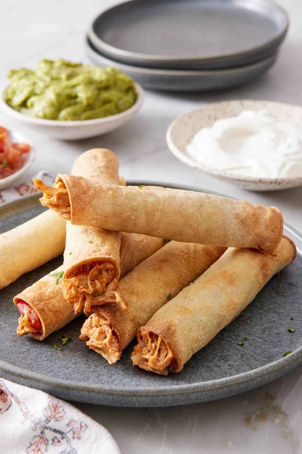 A plate of a pile of air fryer taquitos. Sour cream, guacamole, and plates in the background.