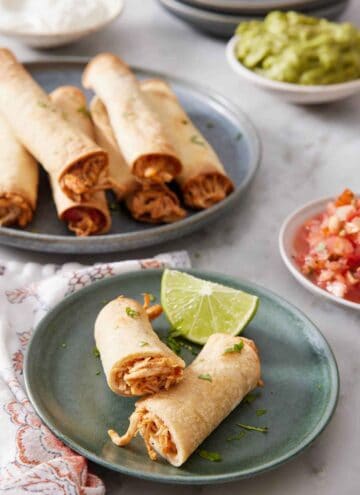 An air fryer taquito cut in half on a green plate with a lime wedge. More taquitos in the background.