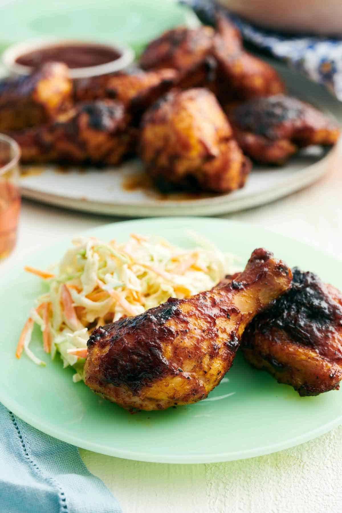 A plate with two pieces of BBQ chicken with coleslaw. A platter in the background with more chicken.