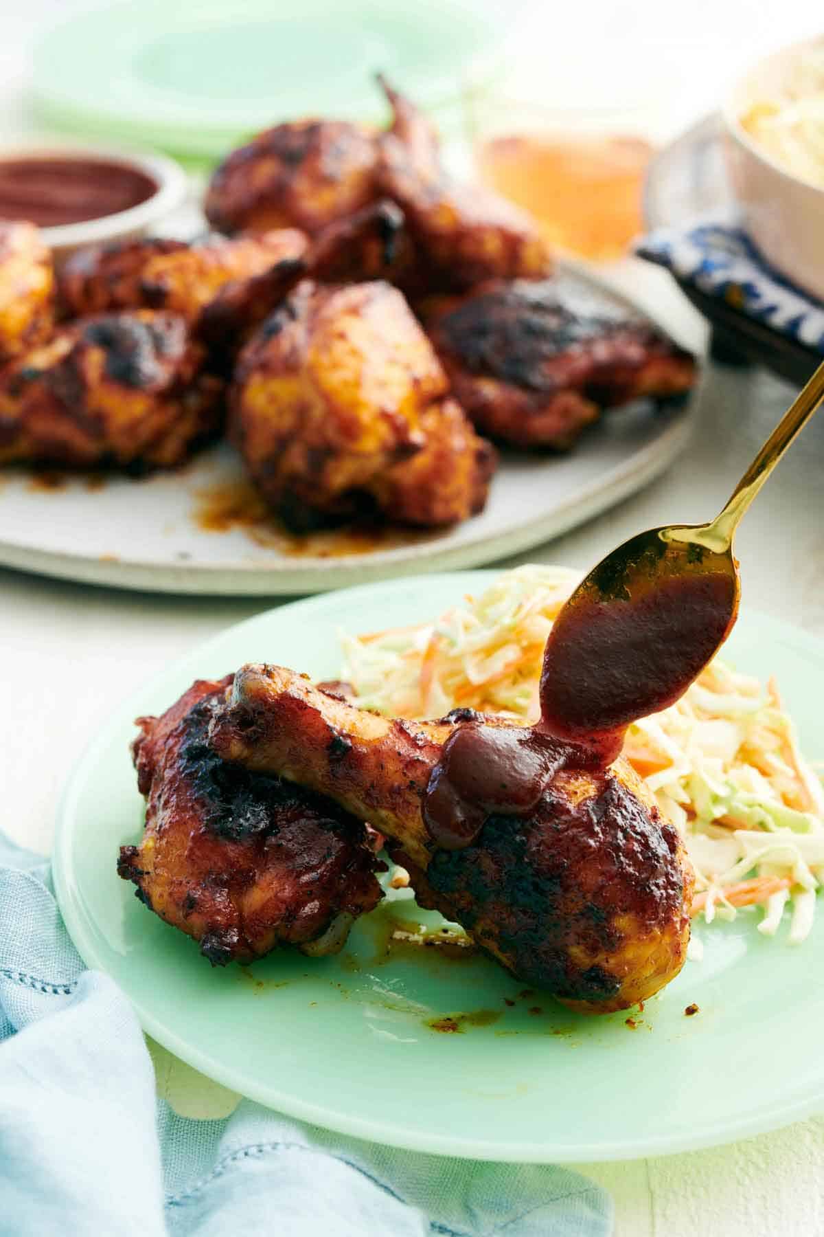 BBQ sauce spooned over a piece of BBQ chicken on a plate with coleslaw. More BBQ chicken in the back on a platter.