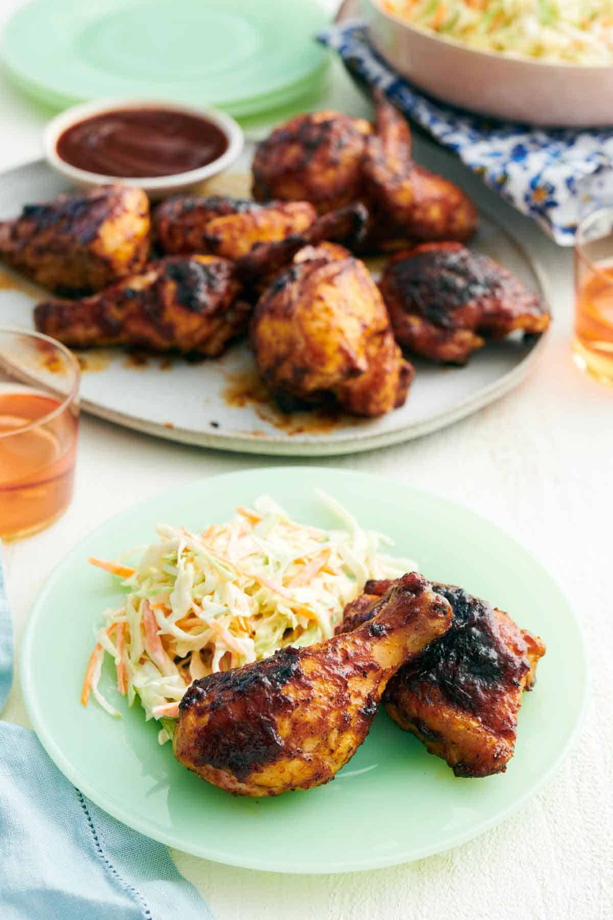 A plate with two pieces of BBQ chicken and coleslaw with a platter of more BBQ chicken in the background.