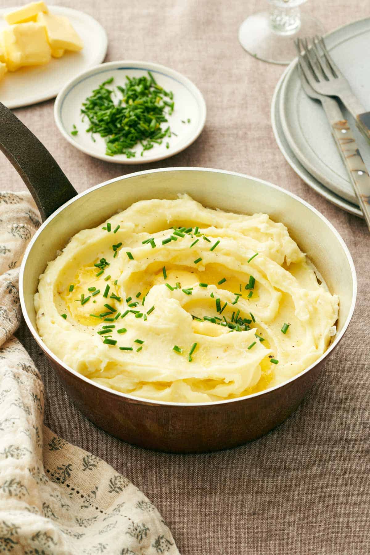 A saucepan with garlic mashed potatoes topped with chives and butter. A plate of butter and chives in the background.