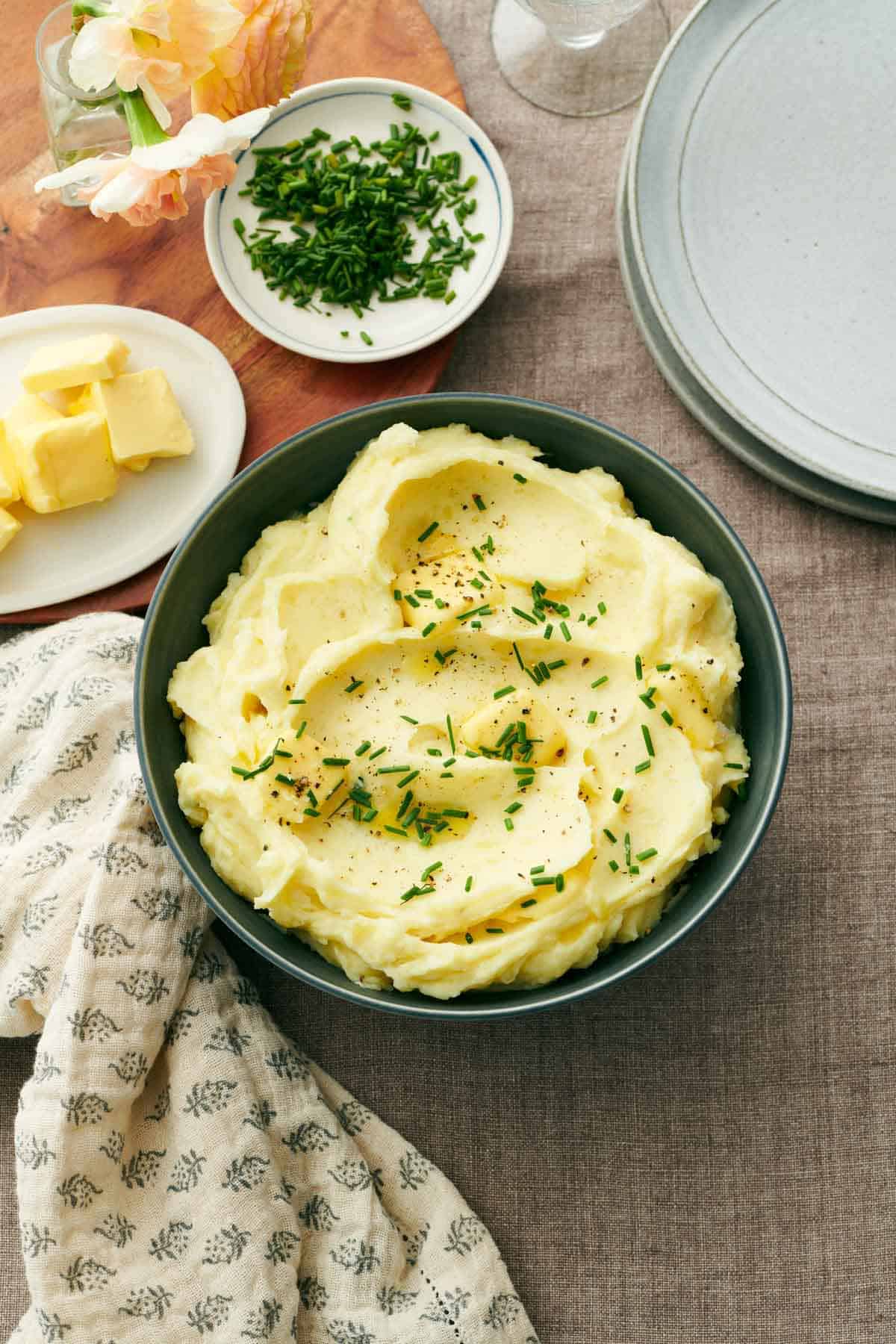 A green bowl of garlic mashed potatoes topped with chives and pepper. A plate of chives, butter, and stack of plates in the back.