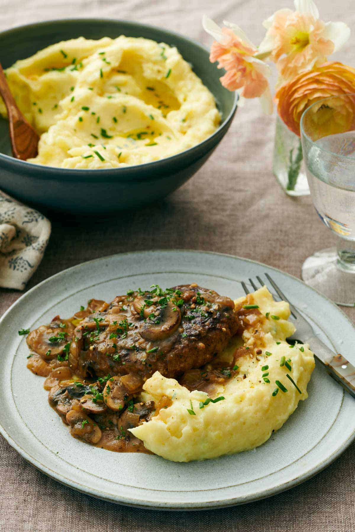 A plate with garlic mashed potatoes with some meat with mushrooms and gravy. A serving bowl of more potatoes in the background.