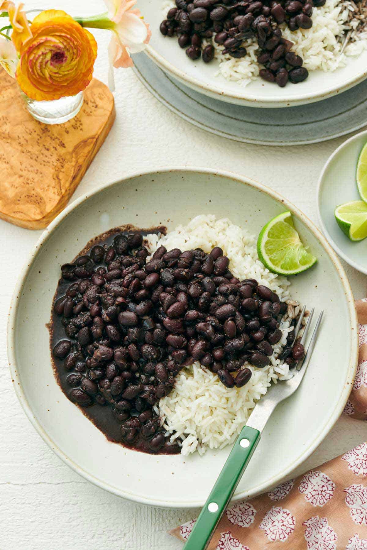 Overhead view of no-soak Instant Pot black beans over rice on a plate with a fork. A second plate with more off to the side and a vase with flowers.