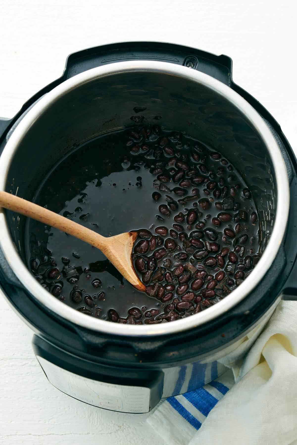 A slightly overhead view of black beans inside of an Instant Pot with a wooden spoon.