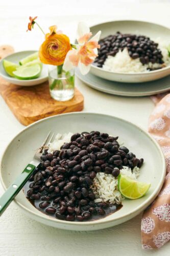 Two plates of Instant Pot black beans over rice with a lime wedge. A vase of flowers and bowl of lime wedges in-between the plates.